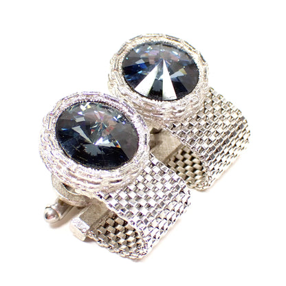 Angled view of the Dante Mid Century vintage rivoli rhinestone cufflinks. The metal is silver tone in color. The tops are round and there is woven mesh chain that wraps around to the back part of the cufflinks. The fronts have faceted country blue rhinestones that come outward to a point.