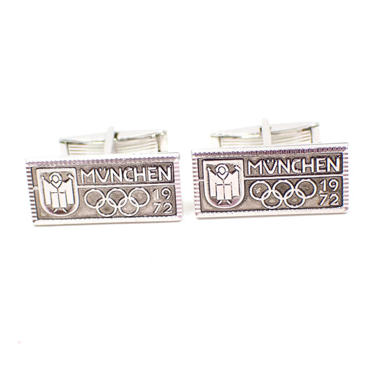 Front view of the 1970's vintage Olympics cufflinks. They are rectangle in shape with a textured edge. There is a person symbol on the left, MÜNCHEN on the top right with a bar underneath it, and then the 5 rings and 1972.