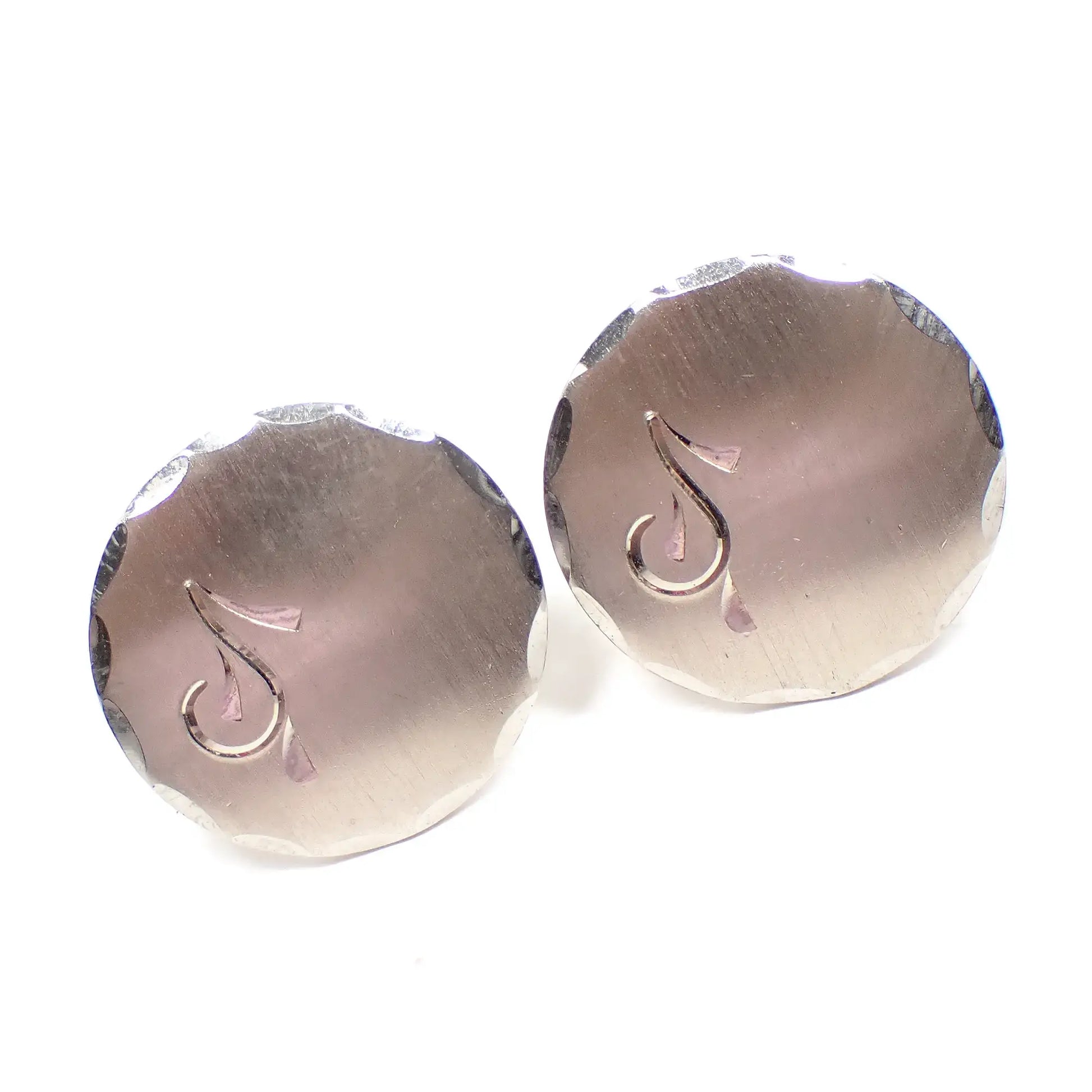 Angled front view of the Mid Century vintage Foster cufflinks. The metal is silver tone in color with a matte brushed appearance on the fronts. They are round shaped with a faceted edge and have an etched design on one side that looks kind of like a musical note.