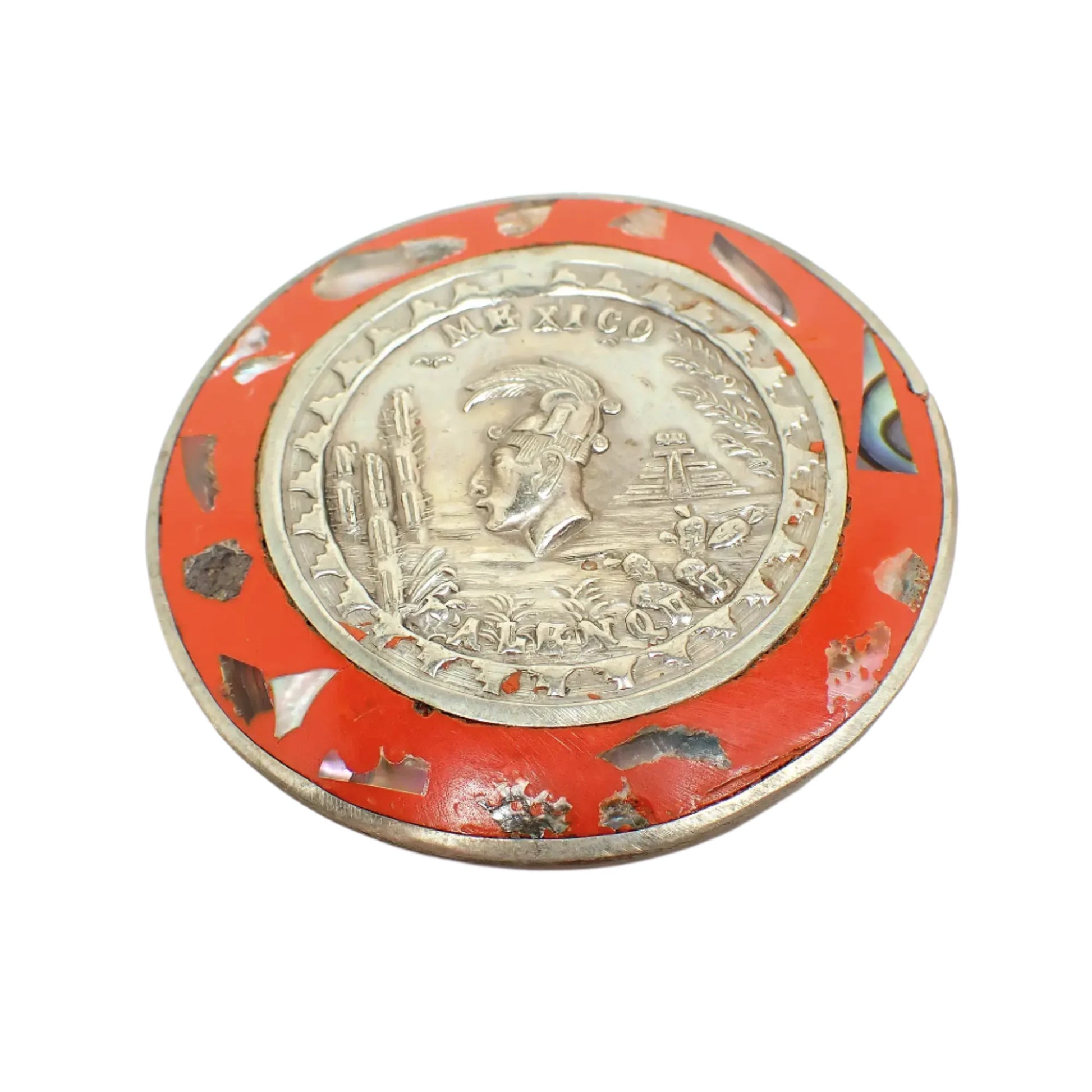 Front view of the brooch. It is round with silver tone metal and dark orange enamel around the edge. There are small pieces of inlaid abalone shell in the enameled area. The middle has a raised depiction of ruins, cacti, and Pakal the Great.