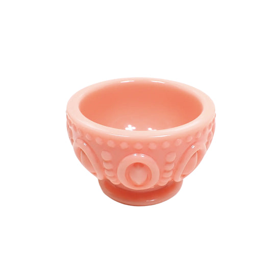 Angled side view of the pastel peach resin footed bowl. It is round with tapered bottom above a flared round footed base. The outside of the bowl has a large raised oval pattern that goes all the way around it. There are lines of raised dots above, below, and in between the ovals. The resin is opaque and shiny in a light pastel peach color.