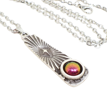 Enlarged close up of the pendant. It is antiqued silver in color and is bar shaped with a flared bottom. The top has an atomic star with radiating rays of light design. On the bottom is a domed round resin cab with matte metallic shades of pink, orange, green, and yellow depending on how the light hits it.