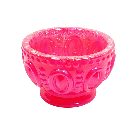 Angled side view of the handmade resin footed bowl. It has bright pearly pink resin with iridescent glitter around the top. It's round and has an oval and dotted design.