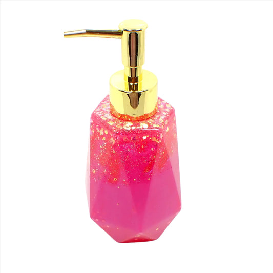 Side view of the handmade resin soap dispenser. In this photo it has a gold tone color pump on it. It is bright pearly pink in color with chunky iridescent glitter mainly on the upper portion of the piece. It has a faceted shape that is narrower at the top and flared at the bottom.
