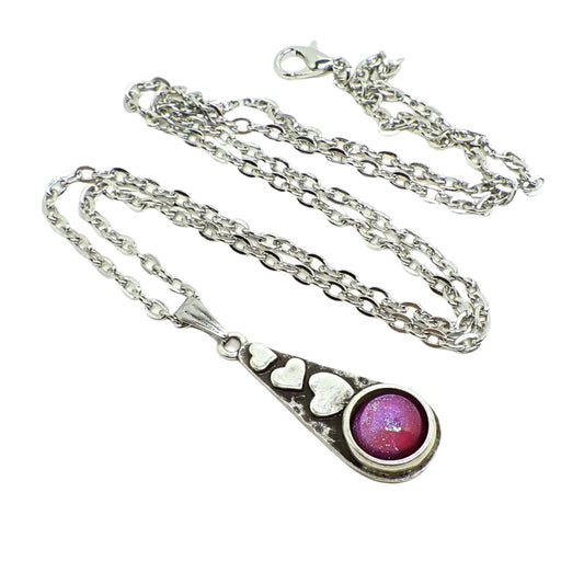 Full view of the heart pendant necklace with pink to purple color shift resin cab. The metal is antiqued silver in color. The necklace has a teardrop shaped pendant with three hearts at the top in a line and and domed round resin cab at the bottom. The cab has color shift resin that goes from pink to purple as the light hits it.