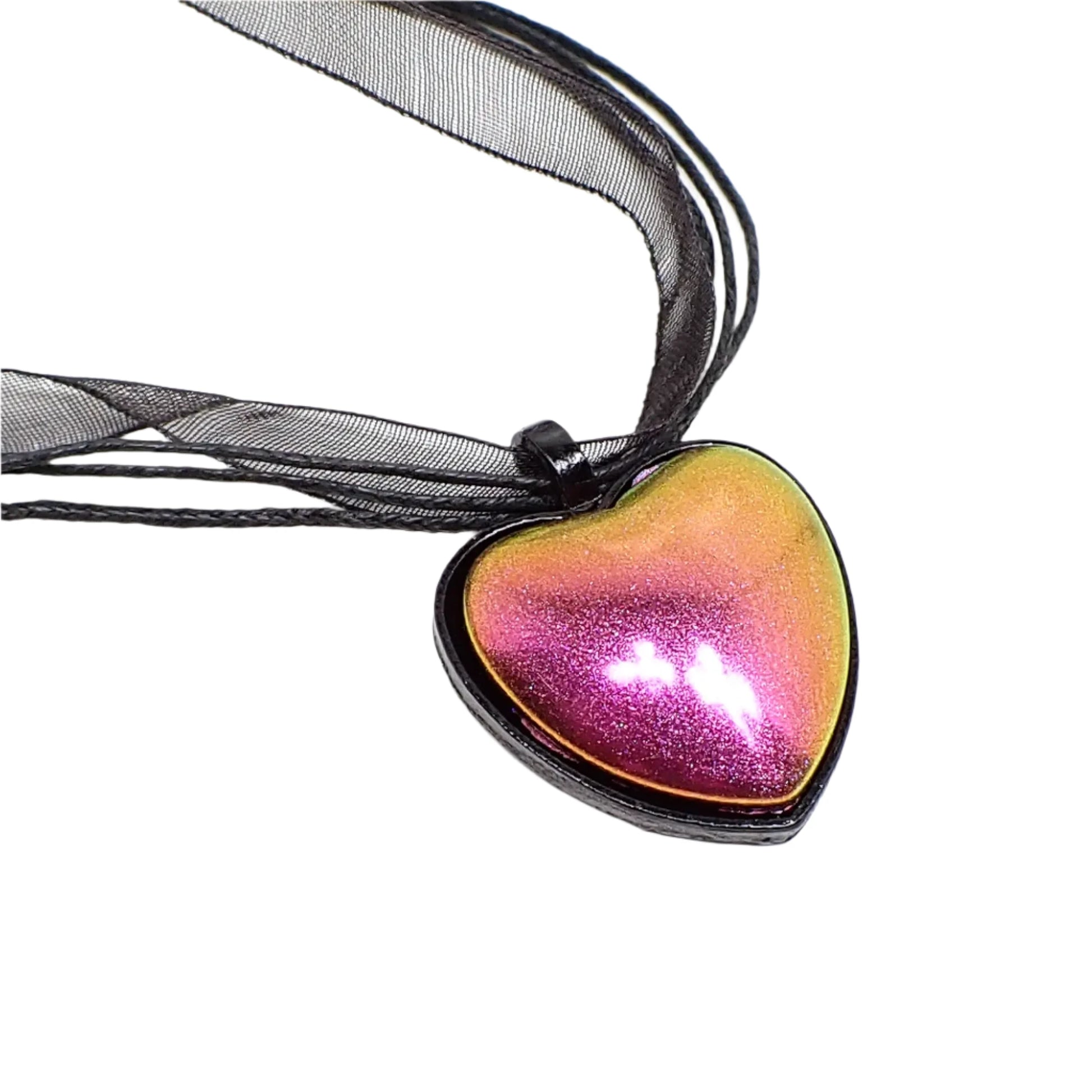 Enlarged Close up photo of the pendant. The setting is black enameled in a heart shape with a loop at the top. The resin cab has shades of bright metallic pink, orange, and yellow showing depending on how the light is hitting it. Organza and strands of waxed cord can be seen going through the top loop.