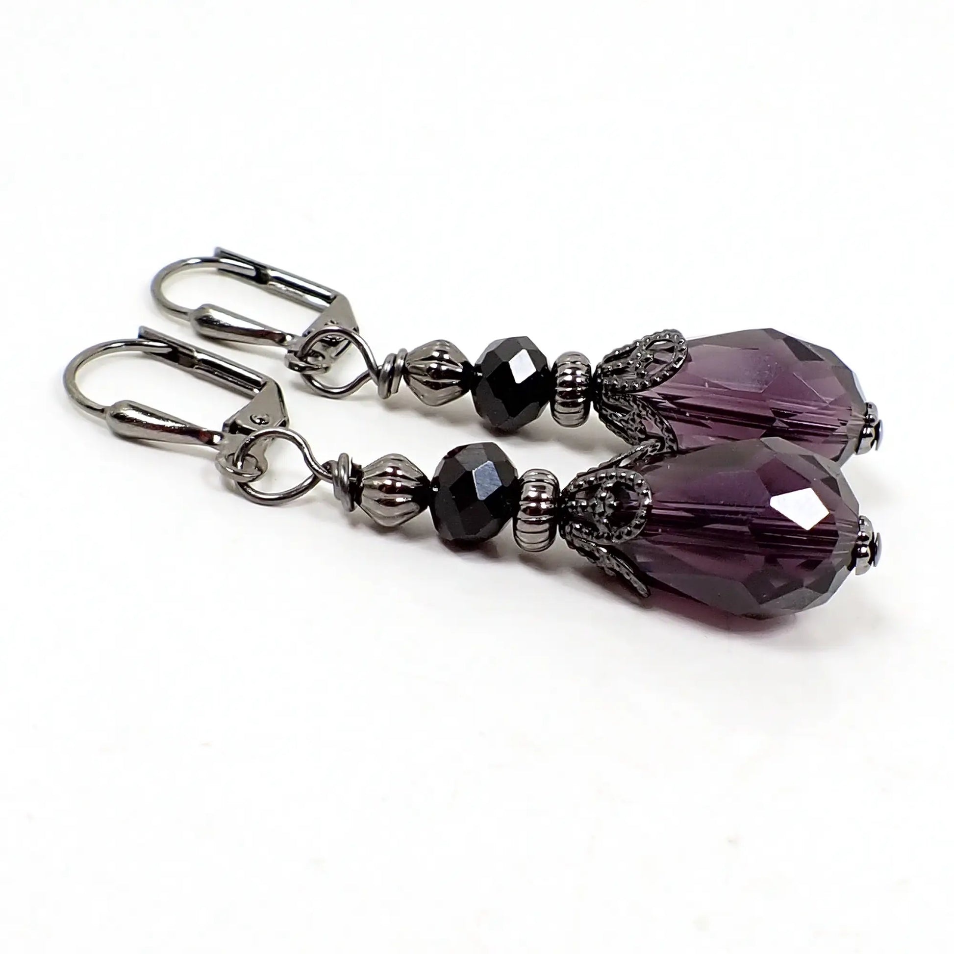 Angled view of the handmade drop earrings. The metal is gunmetal dark gray in color. There is a faceted black glass crystal saucer bead at the top and a faceted dark purple glass crystal teardrop bead at the bottom. 