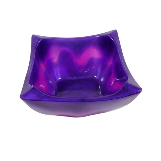 Angled view of the handmade resin retro atomic style square bowl. It has a square shape with curved sides and corners that are angled and taper outwards and upwards. The resin is mostly a vibrant pearly purple color with shades of blue. Semi translucent neon purple resin is marbled into the design around the middle, with some shades of bright pink showing as well. Great for adding a pop of color to your home decor.