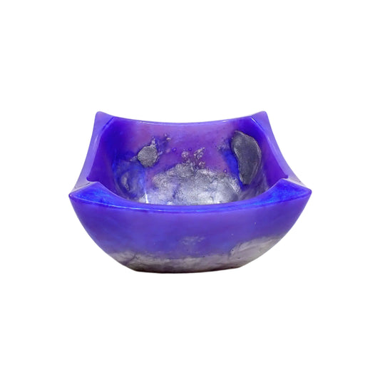 Angled side view of the handmade Retro Atomic style square decorative bowl. It is square with edges that flare outward and upward and have pinched tops. The bottom of the bowl is tapered. The resin is shiny and opaque with marbled swirls of pearly purple and blue with areas of metallic silver on the inside and towards bottom of the bowl.