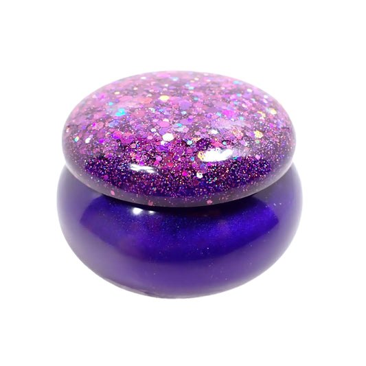 Side view of the handmade resin small round trinket box jar with lid. It has vivid pearly purple resin on the bottom that has flashes of blue color depending on how the light is hitting it. The lid has chunky glitter that has sparkly shades of purple and pink.