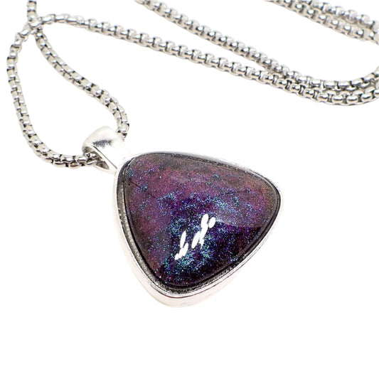 Enlarged close up of the pendant. It has an antiqued silver tone setting in a rounded triangle shape. The domed resin cab has shades of pearly dark purple and blue depending on how the light is hitting it.