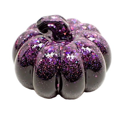 Angled view of the small resin pumpkin. It has shiny black resin with sparkly purple iridescent chunky glitter at the top and stem part of the pumpkin.