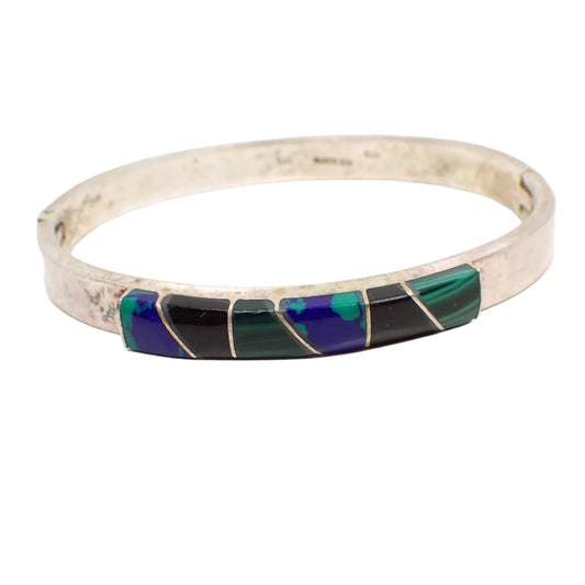 Front view of the vintage sterling silver hinged bangle bracelet with gemstones. The front of the bracelet has geometric shaped slices of gemstone inlaid over resin. There are two each of blue and green color azurite malachite, marbled green malachite, and black onyx gemstone areas. The rest of the bracelet is silver with some darkening of areas from age.