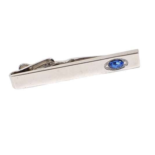 Angled front view of the vintage Swank tie clip. The metal is shiny silver in color. It is bar shaped with a textured oval indent on the end that holds a blue oval rhinestone. 