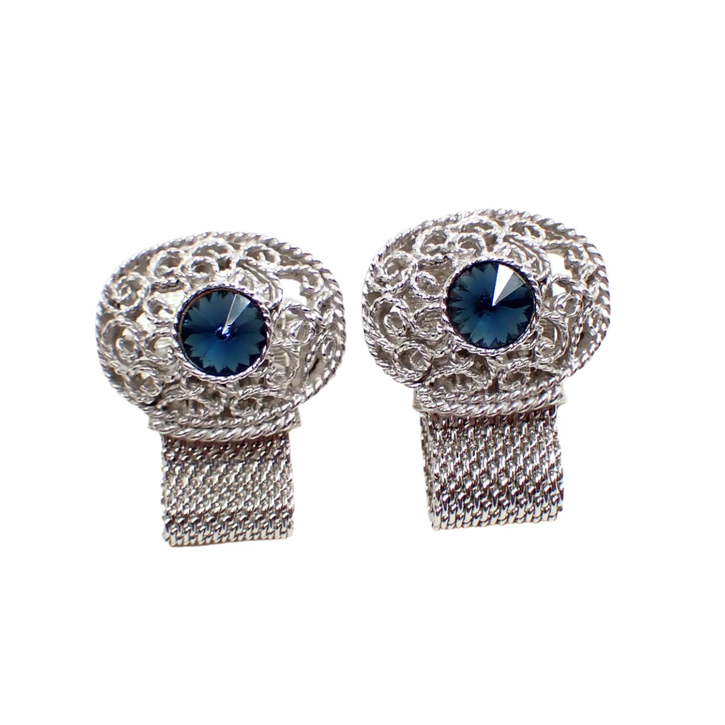 Front view of the Mid Century vintage Swank wrap around cufflinks. The metal is silver tone in color. The tops have large filigree ovals with a denim blue rivoli rhinestone in the middle. There is mesh metal going from the bottom of the ovals around to the back of the cuff links.