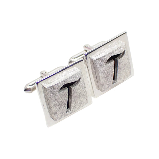 Angled view of the Mid Century vintage Swank cufflinks. The metal is silver tone in color. The fronts have matte brushed silver tone metal with a cut out initial letter T design. 