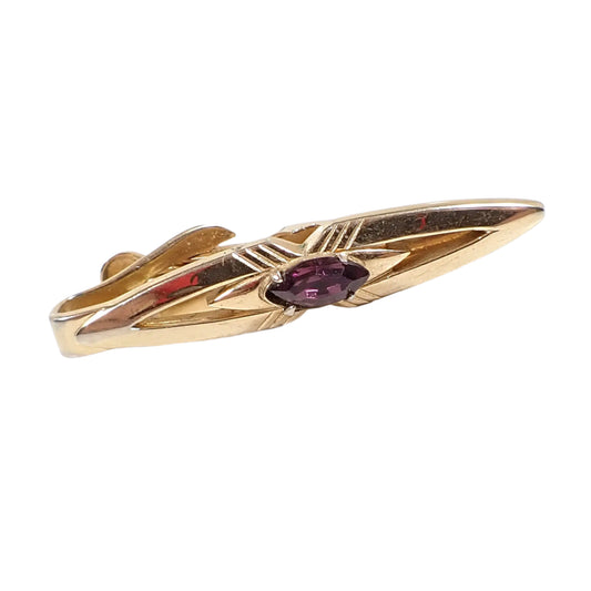 Angled view of the Swank Mid Century vintage tie clip. It has a long marquis shape with a cut out design and engraved lines forming an X in the middle. There is a prong set marquis shaped dark purple rhinestone in the very middle. The metal is gold tone in color.