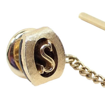 Enlarged view of the vintage Swank initial tie tack. The metal is matte gold tone in color with small lines for a brushed look. It's shaped like a curved rectangle with rounded edges. The middle has a cut out area with the letter S in the center.