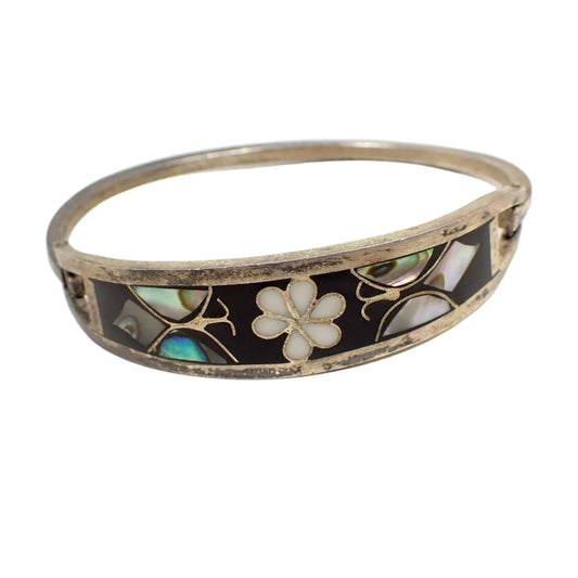 Front view of the vintage Taxco hinged bangle bracelet. The metal has a darkened patina to a dark gray in color. The front is enameled black with a white enameled flower in the middle. There is a butterfly on each side that has inlaid pieces of abalone shell.