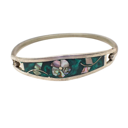 Front view of the vintage Taxco hinged bangle bracelet. The metal is silver tone in color. There is a flower with leaves on either side made of inlaid abalone shell. The flower design is surrounded by pearly green resin enamel.