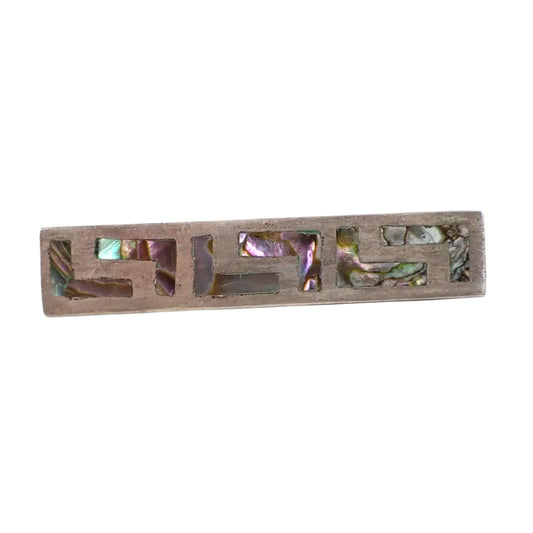 Front view of the vintage Taxco sterling silver and abalone tie bar. The sterling has a darkened patina to an antiqued silver color. The tie clip is shaped like a long rectangle with up and down L like shapes with pieces of inlaid abalone shell in them. The shell pieces are iridescent and have flashes of different colors.