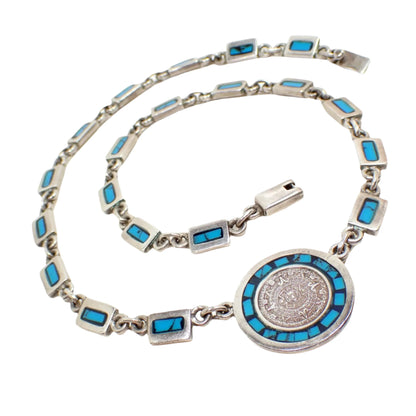 Photo of the Taxco vintage Mayan Calendar necklace. It has sterling silver rectangle links with inlaid blue turquoise chips that are enameled with resin over them. There is a round pendant at the bottom with a raised detailed depiction of a Mayan Calendar. There is a box clasp at the end.