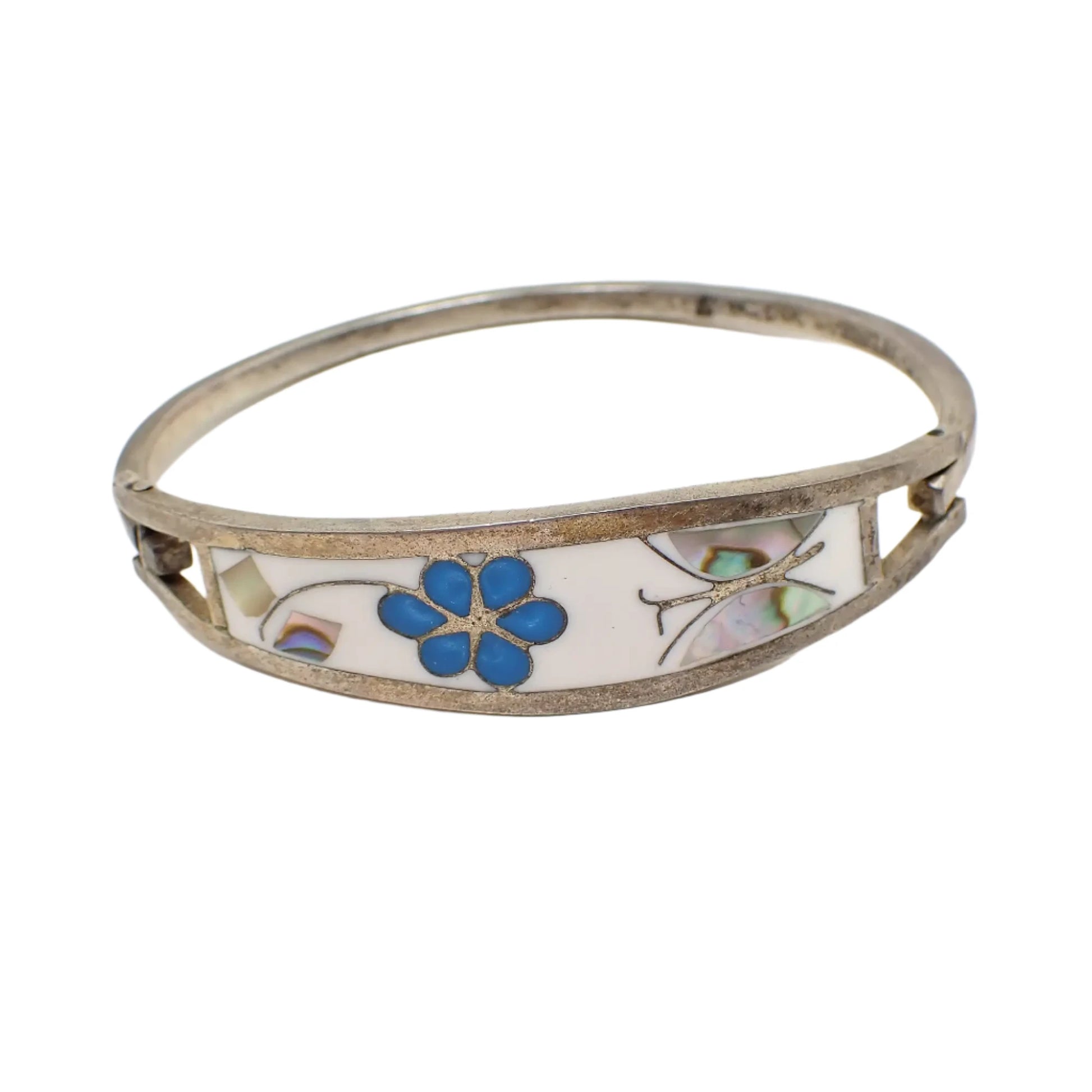 Front view of the Taxco vintage hinged bangle bracelet. The metal is antiqued silver in color. There is a blue enameled flower in the middle surrounded by a white enameled background. On the left side are pieces of abalone shell forming the flower leaves and on the right there are pieces of abalone forming a butterfly.