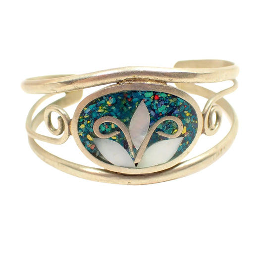 Front view of the vintage cuff bracelet. The metal is silver tone in color. There is an oval on the front with tiny dyed stone chips. The stone chips are different colors, but the piece has an overall teal appearance. There are three pieces of inlaid mother of pearl shell in the middle forming a flower design.