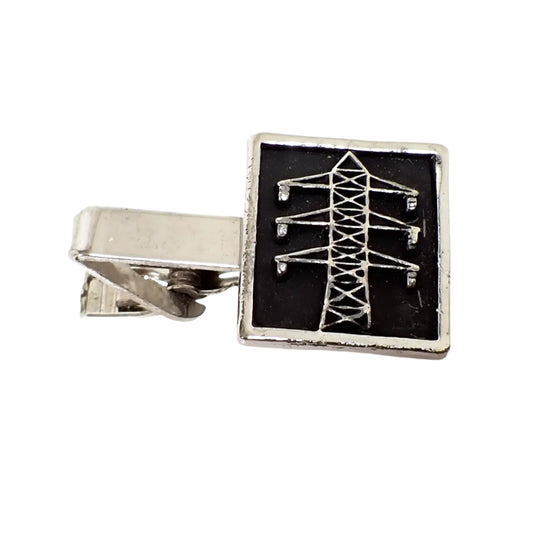 Angled front view of the vintage tie clip. The metal is silver tone in color. There is a raised depiction of an electricity transmission tower on a black painted background in a larger sized rectangle at the end of the tie clip.