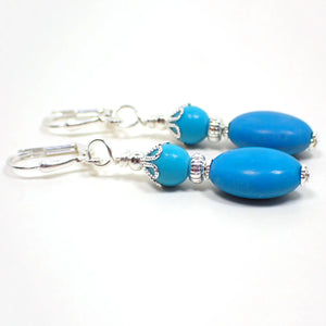 Side view of the handmade magnesite and chalk turquoise drop earrings. The metal is silver plated in color. There are small round blue dyed magnesite beads at the top. The bottom beads are puffy oval shaped and are chalk turquoise which is ground down, dyed, and then added to resin to form the beads.