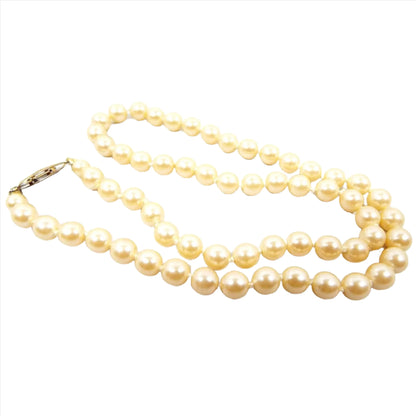 Angled view of the Mid Century vintage faux pearl beaded necklace. The glass imitation pearls are a golden off white in color. There is a gold tone plated slide clasp at the end.