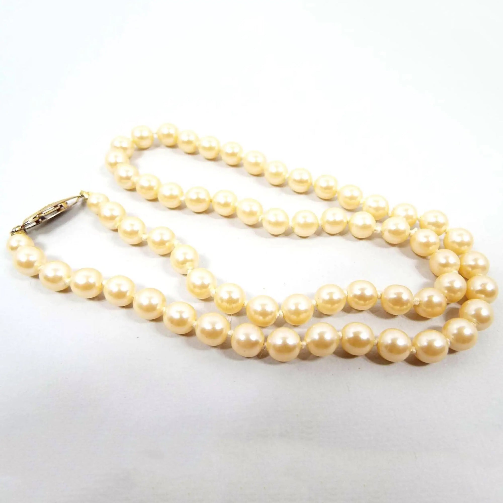 Angled view of the Mid Century vintage faux pearl beaded necklace. The glass imitation pearls are a golden off white in color. There is a gold tone plated slide clasp at the end.