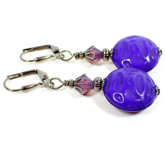 Side view of the handmade drop earrings. The metal is gunmetal gray in color. There are light purple faceted glass crystal beads at the top with hints of pink. The bottom vintage beads are lucite, are puffy round in shape, and have a textured indented pattern on them. They are a vivid bluish purple in color.