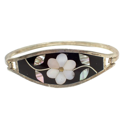 Front view of the vintage Alpaca Mexico hinged bangle bracelet. It has silver tone color metal. There is a hinge on one side and clasp on the other. The front is curved and has black enamel with inlaid pieces of mother of pearl and abalone shell making up a floral design. The mother of pearl is the flower in the middle and the abalone makes up the two leaves on stems on either side.