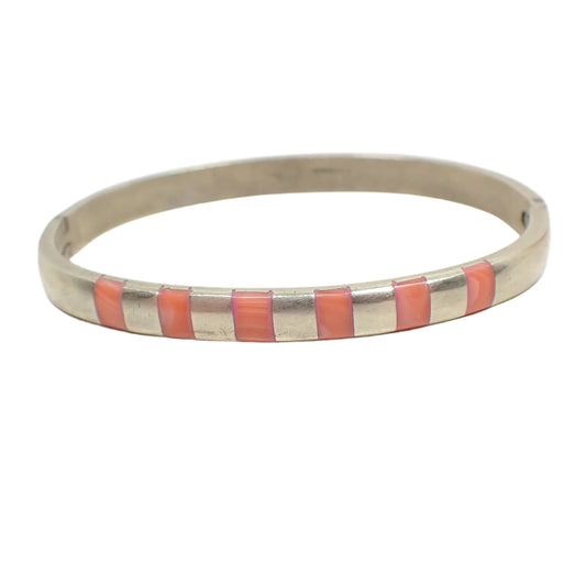 Front view of the Mexican vintage hinged bangle bracelet. It has a slightly darkened patina from age. The bracelet is oval with a curved band of sterling silver that has rounded edges. The front has six pieces of inlaid salmon pink colored coral secured with resin. 