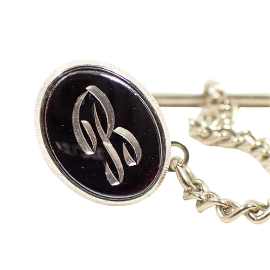 Front view of the Mid Century vintage faux hematite tie tack. The metal is silver tone in color. The tie tack is oval and has a dark metallic gray imitation hemtatite front with a fancy script initial B in the middle.