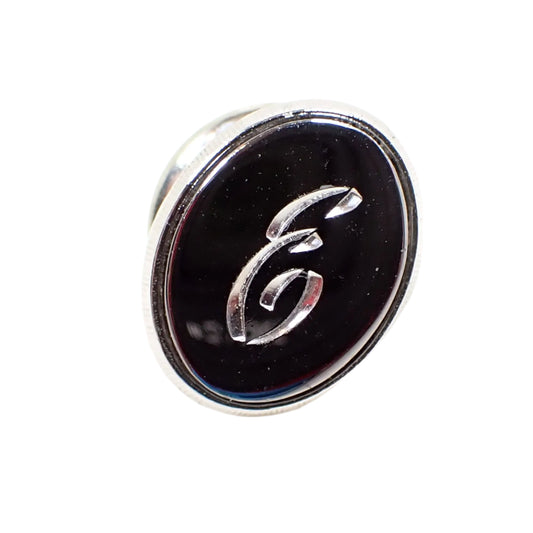 Front view of the Mid Century vintage faux hematite tie tack. The metal is silver tone in color. The tie tack is oval and has a dark metallic gray imitation hemtatite front with a fancy script initial E in the middle. 