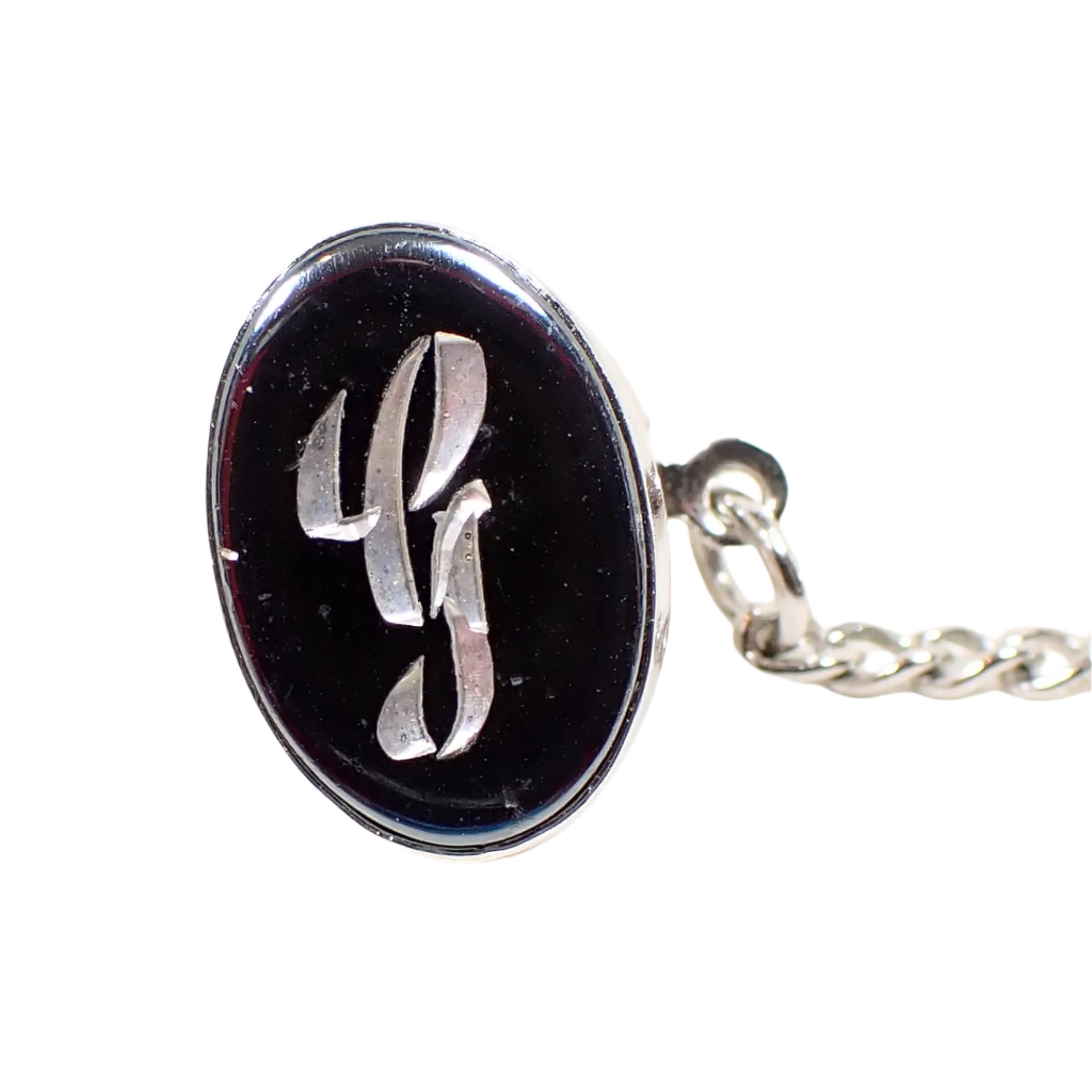 Front view of the Mid Century vintage faux hematite tie tack. The metal is silver tone in color. The tie tack is oval and has a dark metallic gray imitation hemtatite front with a fancy script initial G in the middle.
