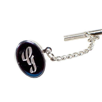 Full view of the vintage initial tie tack. It is oval shaped with silver tone color metal and a dark metallic gray faux hematite front. There is a fancy script letter G engraved in the middle.