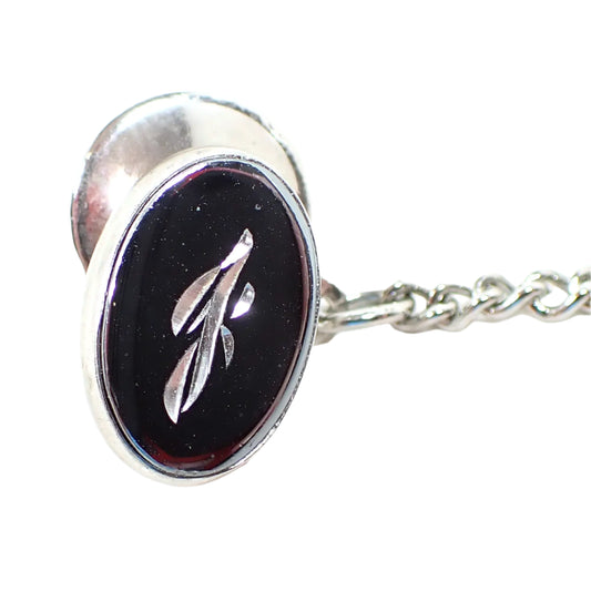 Front view of the Mid Century vintage faux hematite tie tack. The metal is silver tone in color. The tie tack is oval and has a dark metallic gray imitation hemtatite front with a fancy script initial J in the middle.