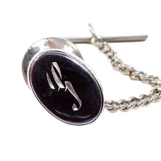 Front view of the Mid Century vintage faux hematite tie tack. The metal is silver tone in color. The tie tack is oval and has a dark metallic gray imitation hemtatite front with a fancy cursive script initial S in the middle.