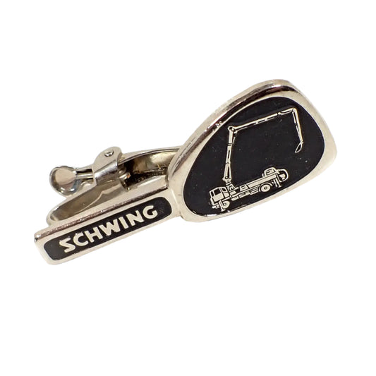 Angled view of the front of the tie clip. The metal is silver tone in color. The rectangle bar part on the left has "Schwing" on a black painted background. The end of the tie clip has a rounded asymmetrical shape with a raised silver tone depiction of a Schwing Roll and Fold Boom concrete pump on a black painted background.