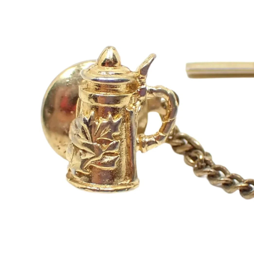 Enlarged view of the vintage stein tie tack. The metal is gold tone in color. It is shaped like a an old stein and has a raised design with leaves on the front.