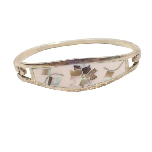 Angled front view of the Mexican vintage hinged bangle bracelet. It has silver tone color metal with a hinge on the side. The front has white enamel with inlaid pieces of abalone shell in a flower and leaves design. 