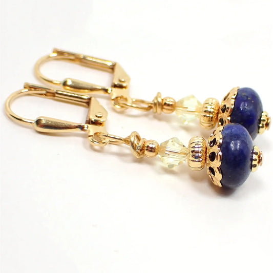 Angled view of the small handmade lapis lazuli drop earrings. The metal is gold plated in color. There are light yellow faceted glass crystal beads at the top. The bottom gemstone beads are a dark blue in color with tiny flecks of gold color. The beads are rondelle saucer like shaped.