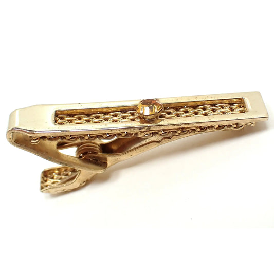 Side and top view of the 1960s Mid Century vintage Hickok tie clip. The metal is gold tone plated in color. It has a rectangle design with open middle chain mesh area. There is an orange rhinestone in the middle. 