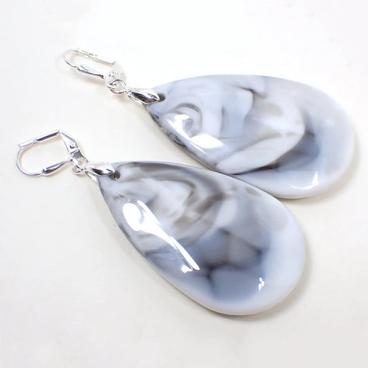 Angled view of the big handmade acrylic teardrop earrings. The metal is silver tone plated in color. They are large puffy teardrop shaped acrylic that are mostly white with gray and some small areas of black marbled in.