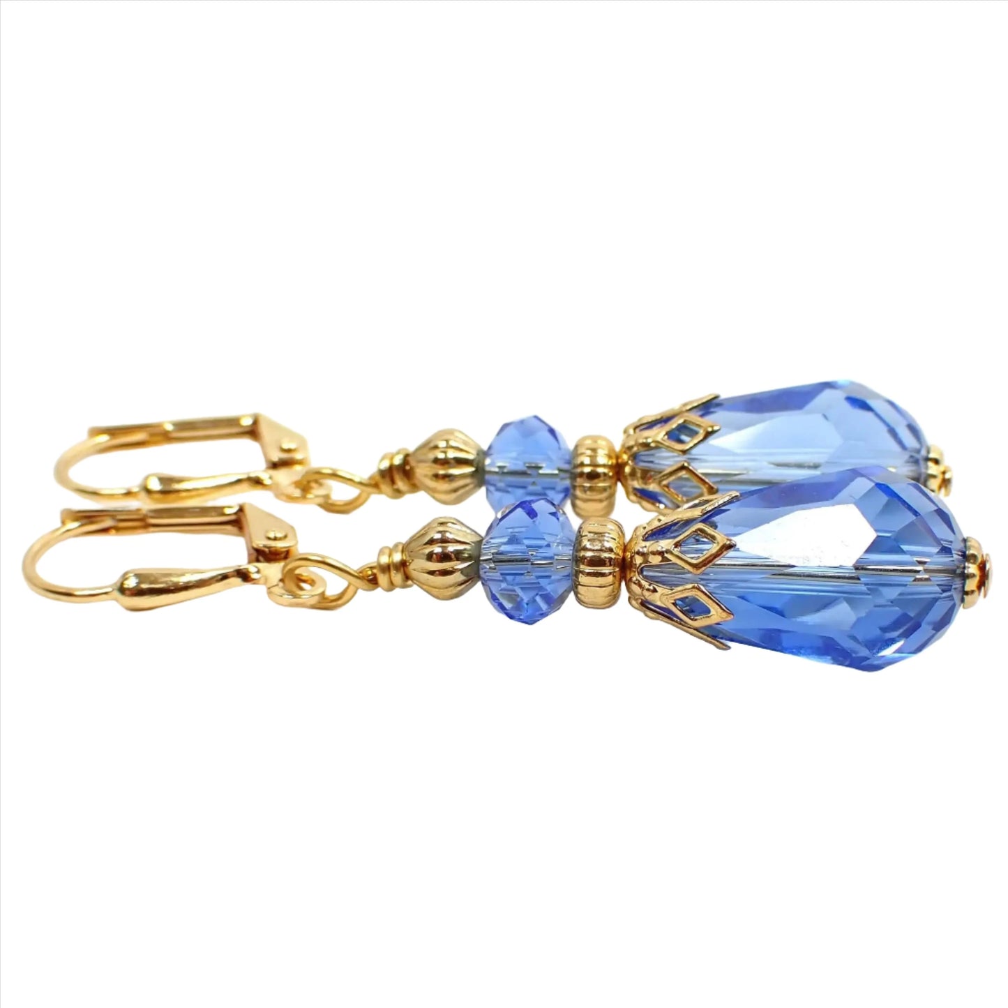 Side view of the handmade glass crystal teardrop earrings. The metal is gold tone in color. There is a cornflower blue faceted oval glass crystal bead at the top and a cornflower blue faceted glass crystal teardrop bead at the bottom.