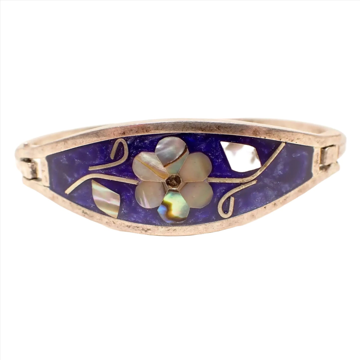Front view of the retro vintage Mexican floral hinged bangle bracelet. The metal is slightly darkened silver tone in color. There is pearly dark blue resin enamel on the front that is like a purplish blue color. The flower in the middle has petals and leaves of inlaid abalone shell.