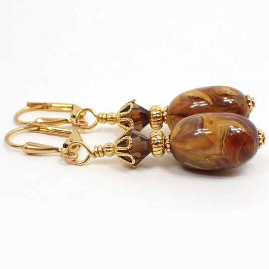 Side view of the handmade oval drop earrings made with vintage lucite beads. The metal is gold plated in color. There are new brown faceted glass crystal beads at the top. The bottom lucite beads have marbled swirls of mostly brown and yellow color with some orange here and there. Each lucite bead is different in pattern and color swirls.